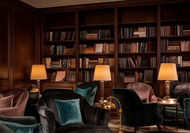 Lounge interna del club con poltrone in velluto scuro, lampade da lettura e libreria in legno scuro su parete